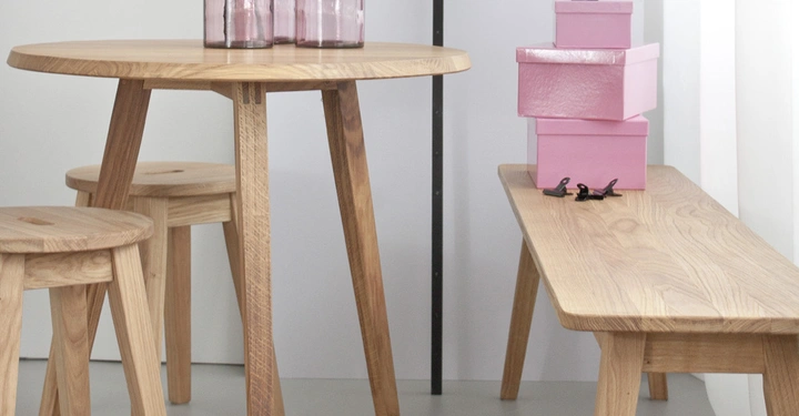 Galerie-Bild 2: Drei rosa Glasflaschen stehen auf einem runden Holztisch in einem hellen Raum mit weißen Wänden. Daneben befindet sich eine Holzbank, auf der drei pinke Kisten gestapelt sind.