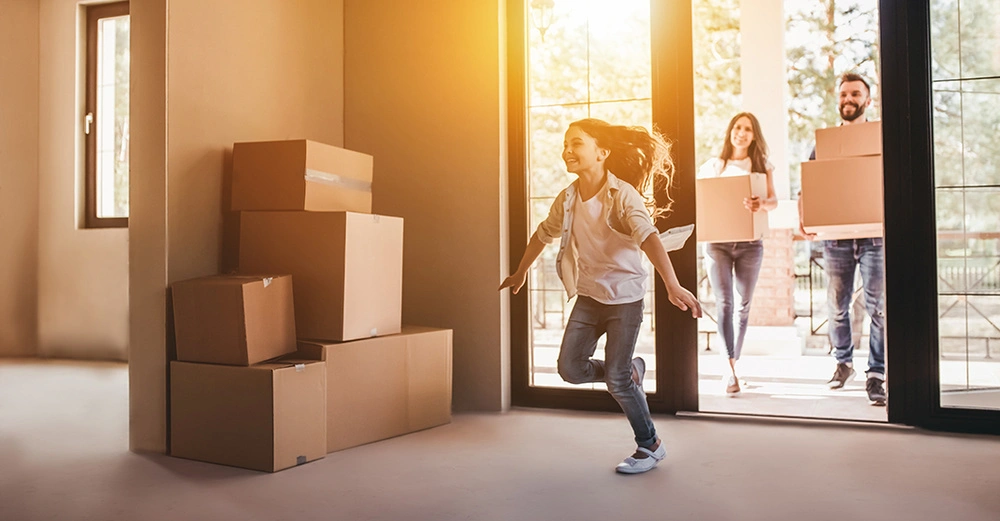 Familie betritt neues Haus mit Umzugskartons. Fröhliches Mädchen läuft vor und warmes Sonnenlicht fällt durch die Fenster.