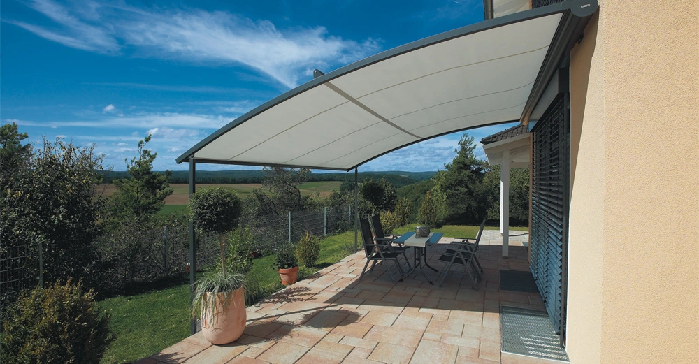Überdachte Terrasse mit Tisch und Stühlen im Garten neben einem Haus in ländlicher Umgebung mit freiem Blick auf grüne Landschaft und einen hellblauen Himmel mit leichter Bewölkung.