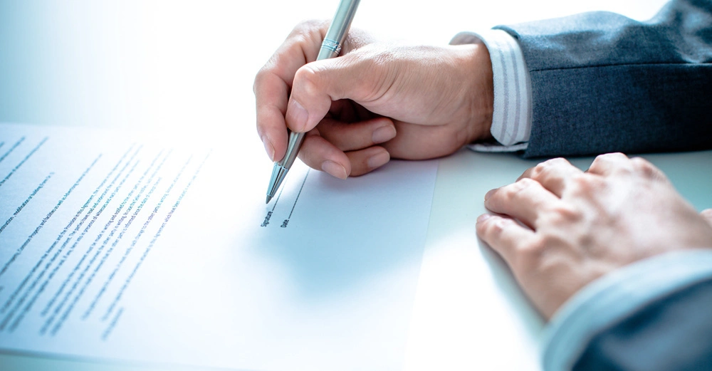 Eine Hand in einem grauen Anzug schreibt mit einem Stift auf ein Dokument auf einem weißen Tisch. Text steht auf dem Dokument: Signature Date