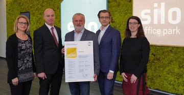 Fünf Personen stehen in einem Bürogebäude vor zwei Schildern mit der Aufschrift silo office park Eine Person hält ein DGNB-Zertifikat mit dem Text Silo Plus.