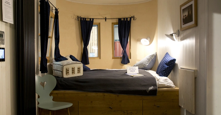 Galerie-Bild 4: Holzbett mit dunkelblauer Bettdecke in kleinem Zimmer mit weißen Wänden dunkelblauen Vorhängen und Schreibtischstuhl darunter Schild mit Aufschrift: Bettenwechsel ohne Leinen 10-12 Uhr