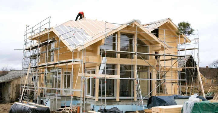 Holzhaus im Bau eingerüstet in ländlicher Umgebung Arbeiter auf dem Dach Stapel von Baumaterialien und abgedeckte Materialien im Vordergrund keine erkennbare Schrift