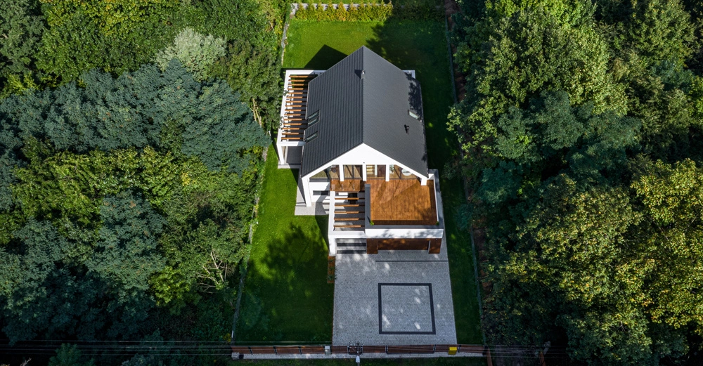 Haus mit schwarzem Dach steht in einem Garten umgeben von dichten Bäumen Terrasse aus Holzplatten vorne und hinten Steinplatten vor dem Haus und grüner Rasen.