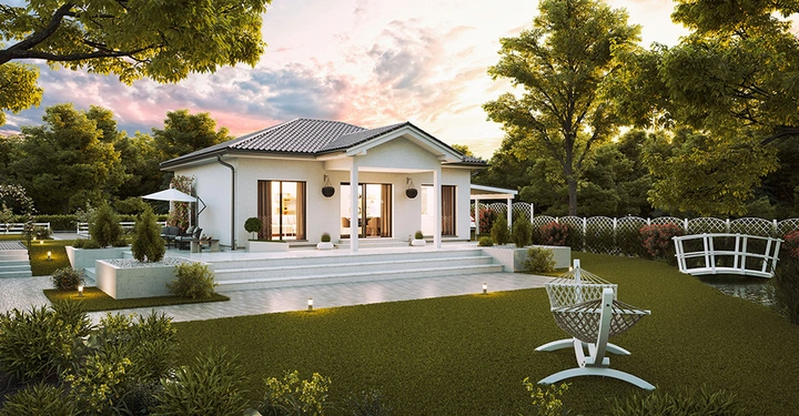 Galerie-Bild 6: Bungalow mit Steildach und großer, mit sehr hellem Stein gepflasterter Terrasse, abendlich beleuchtet, steht in einem großen Garten mit hohen Bäumen und saftigm grünem Rasen. 