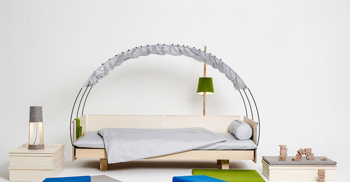 Galerie-Bild 2: Holzbett mit grauem Baldachin und Bettwäsche in modernem, minimalistischem Raumkontext, umgeben von Möbeln, Kissen und Holzbausteinen. Eine grüne Lampe steht rechts, eine graue Lampe links.