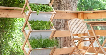 Pflanzen wachsen in einem vertikalen Holzrahmen auf einer Terrasse neben einem Holztisch und einem Stuhl unter einem großen Baum mit üppigem grünen Laub.