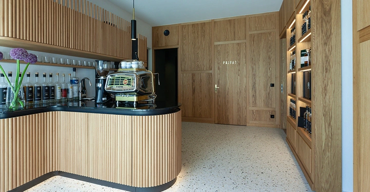 Galerie-Bild 11: Kaffeemaschine auf einer Theke in einer stilvollen Holzverkleidung Küchenschrank mit Flaschen im Hintergrund Holzwände und eine Tür mit der Aufschrift PRIVAT in einem modernen Raum.