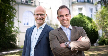 Zwei Männer lächeln in die Kamera einer steht mit verschränkten Armen der andere trägt eine Brille sie befinden sich draußen vor einem Gebäude mit weißer Fassade und großen Bogenfenstern