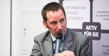 Mann im Anzug spricht in ein Mikrofon sitzend vor einem Plakat auf dem steht INFORMATION SERVICE AUSTAUSCH AKTIV FÜR SIE.