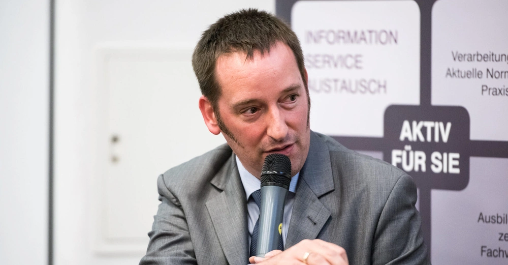 Mann im Anzug spricht in ein Mikrofon sitzend vor einem Plakat auf dem steht INFORMATION SERVICE AUSTAUSCH AKTIV FÜR SIE.