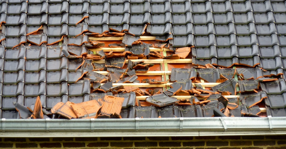 Ziegel auf einem Hausdach sind zerbrochen. Zerbrochene Dachziegel liegen gestapelt. Ein intaktes Regenrinnensystem befindet sich unten. Das restliche Dach ist mit intakten Ziegeln gedeckt.