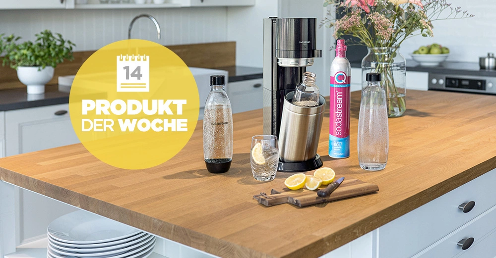Sprudlergerät steht auf einer Holzarbeitsplatte in einer modernen Küche mit weißen Schränken und einem Wasserhahn im Hintergrund Davor liegt eine Zitronenscheibe daneben ein Glas Wasser und eine Karaffe. Text: 14 PRODUKT DER WOCHE