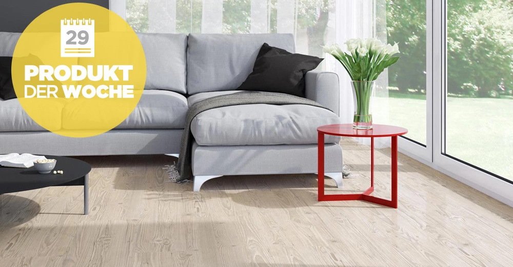 Graues Sofa steht vor großen Fenstern im hellen Wohnzimmer roter Beistelltisch mit weißem Blumenstrauß daneben Text auf gelbem Kreis 29 PRODUKT DER WOCHE