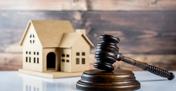 Holzhammer liegt auf Holzblock neben Pappmodell eines Hauses auf einer glatten Oberfläche im Vordergrund Holzverkleidung im Hintergrund