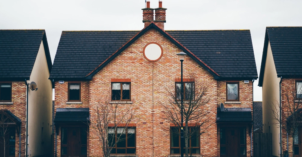 Backsteinhaus mit Spitzdach steht in einer Reihenhaussiedlung zwei kahle Bäume davor Laternenmast in der Mitte der ruhigen Straße. Keine identifizierbaren Personen oder Tiere sichtbar.