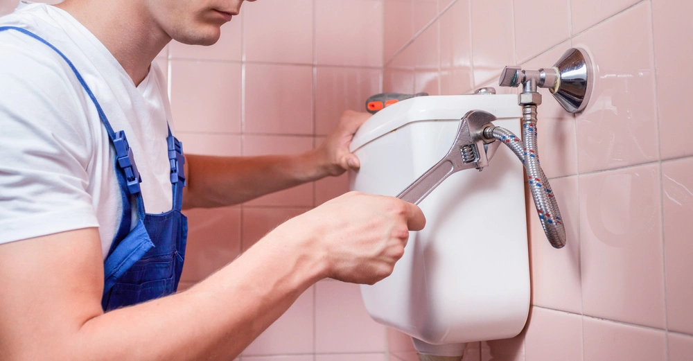 Mann arbeitet mit einem Schraubenschlüssel an einem Toilettenspülkasten in einem Badezimmer mit rosa gefliesten Wänden