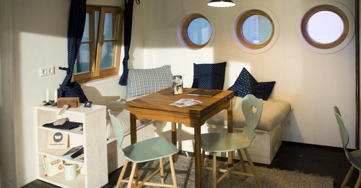 Galerie-Bild 3: Holztisch mit grünen Stühlen steht in einer gemütlichen Sitzecke mit Sofa und Kissen vor rundem Fenster Schatten. Rechts eine Hängeleuchte. Links Regal mit Büchern und Deko. Text: W wohnwagen