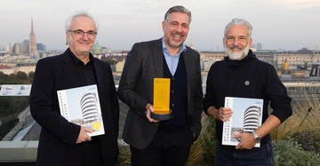 Drei Männer halten Auszeichnungen auf einer Dachterrasse mit Blick auf eine Stadt im Hintergrund. Der mittlere Mann hält eine gelbe Trophäe mit der Aufschrift: Award Wohnbauten des Jahres 2023 Acknowledgement Quadriga Hochschule.