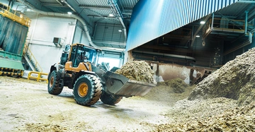 Ein Radlader schaufelt Material in einer großen, gut beleuchteten Industriehalle mit Metallkonstruktionen, Maschinen und Rohren.