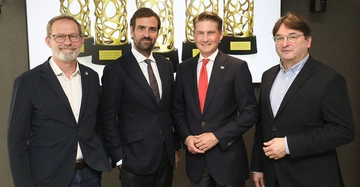 Hannes Horvath, Georg Flödl, Matthias Gass und Anton Holzapfel stehen nebeneinander und lächeln in die Kamera. Alle tragen einen dunklen Anzug. Auf einem Bildschirm im Hintergrund ist die goldene Trophäe des Prix d'Excellence zu sehen.