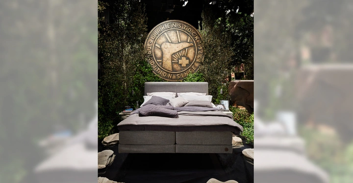 Ein Bett steht in einem Raum mit üppiger grüner Dekoration, auf dem Hintergrund ein großes rundes Wandrelief mit der Aufschrift "BIRKENSTOCK EDITION SINCE 1774"