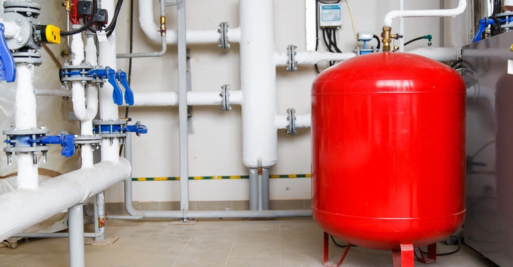 Ein roter Druckbehälter steht auf dem Boden neben isolierten weißen Rohrleitungen mit blauen Ventilen in einem Maschinenraum mit grauen Wänden und Fliesenboden.