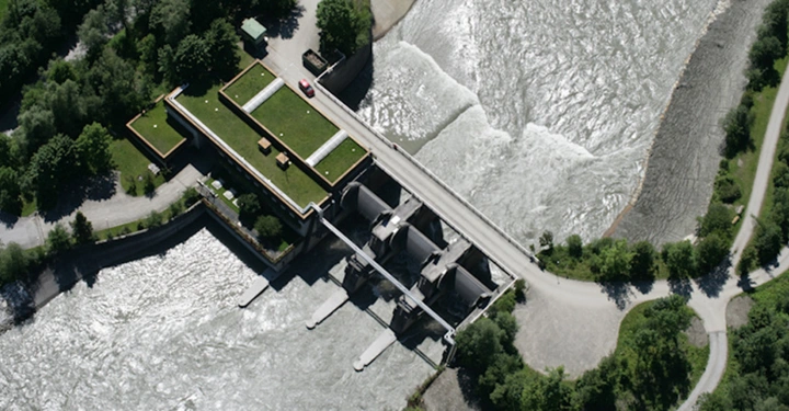 Wasserkraftwerk mit Staumauer und Schleusen steuert den Wasserfluss eines Flusses eingebettet in eine grüne bewaldete Landschaft mit umliegenden Wegen und Straßen. Ein rotes Auto parkt auf der Staumauer.