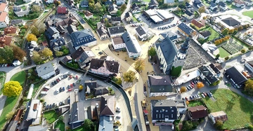 Luftaufnahme einer kleinen Stadt Häuser stehen dicht beieinander Autos sind auf Parkplätzen verteilt Bäume und Grünflächen umgeben Gebäude ein großer Friedhof neben einer Kirche im Zentrum des Asphaltnetzes