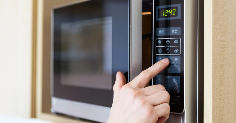 Hand drückt auf die Tasten eines Mikrowellenherds in einer Küchenumgebung Das Display zeigt 12:48 an und die Tasten umfassen verschiedene Symbole für Automatikmenüs und Zeiteinstellungen