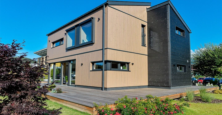 Modernes zweistöckiges Haus mit hellen Holzverkleidungen und schwarzen Akzenten steht auf einer Holzterrasse umgeben von gepflegtem Rasen und Sträuchern unter klarem blauem Himmel in einer Wohngegend