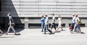 Mehrere Menschen gehen seitlich auf einem Bürgersteig entlang einer grauen Betongebäudewand bei Tageslicht. Einige tragen lässige Kleidung und tragen Taschen oder trinken etwas.