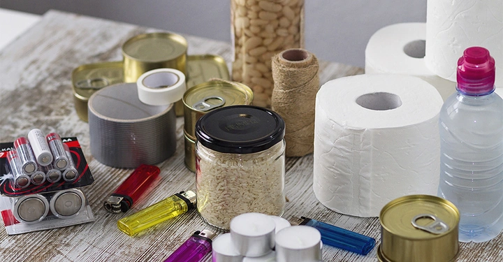 Verschiedene Vorratsgegenstände wie Konservendosen Batterien Reis Toilettenpapier Feuerzeuge Flaschenband und eine Wasserflasche liegen auf einem Holztisch in einem Raum