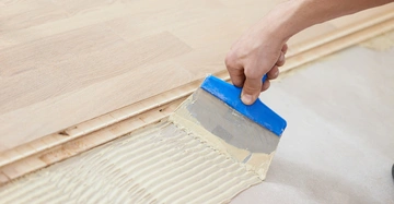 Eine Hand verwendet eine Kelle mit blauem Griff zum Auftragen von Klebstoff auf einen Betonboden im Rahmen der Vorbereitung zur Verlegung von Parkettboden.