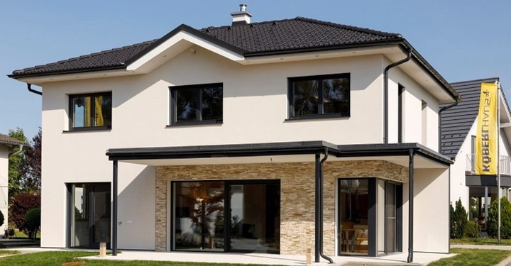 Haus mit zwei Stockwerken und moderner Architektur steht auf einer sauberen Straße bei sonnigem Wetter. Im Hintergrund ist ein gelbes Banner mit der Aufschrift KÖBERL HAUS.