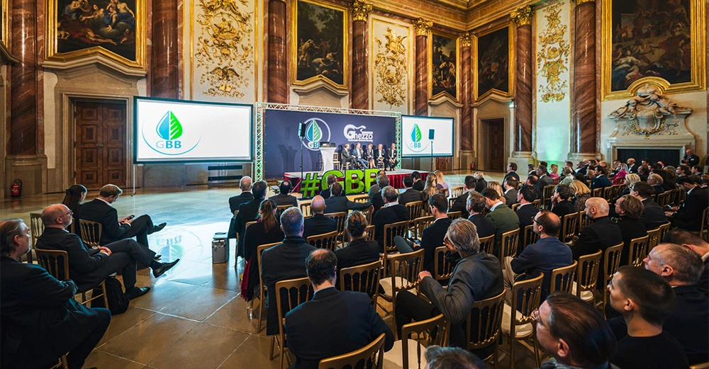 Menschen sitzen und stehen in einem prunkvollen Raum mit Gemälden an den Wänden und hören einer Podiumsdiskussion auf einer Bühne zu. Zwei Bildschirme zeigen das Logo "GBB".