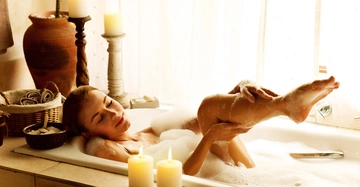 Frau badet in einer Wanne mit Schaum und hebt ihr Bein an Umgeben von Kerzen einem großen Tonkrug und einem Korb mit Handtüchern in einem Raum mit hellen Vorhängen