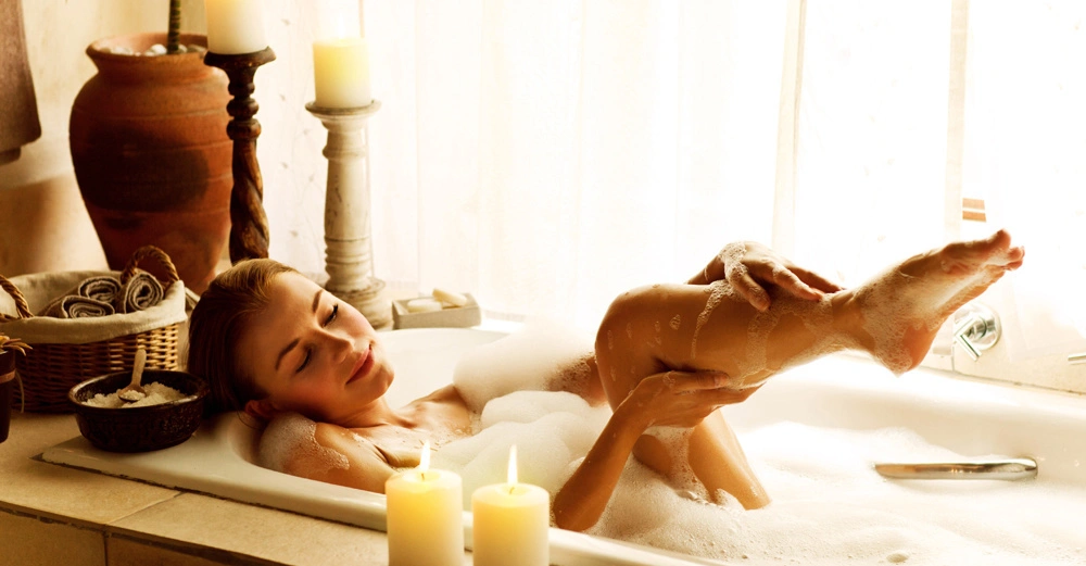 Frau badet in einer Wanne mit Schaum und hebt ihr Bein an Umgeben von Kerzen einem großen Tonkrug und einem Korb mit Handtüchern in einem Raum mit hellen Vorhängen