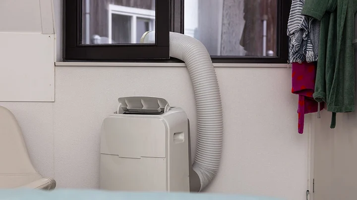 Eine weiße mobile Klimaanlage steht direkt vor einem geöffneten Fenster, durch das der Abluftschlauch die heiße Luft nach draußen leitet.