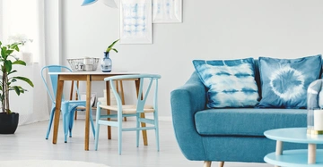 Blaues Sofa mit Kissen steht vor einem Holztisch mit hellblauen Stühlen Ein Metallkorb und eine Vase befinden sich auf dem Tisch Pflanzen und Wandbilder schmücken den hellen Raum