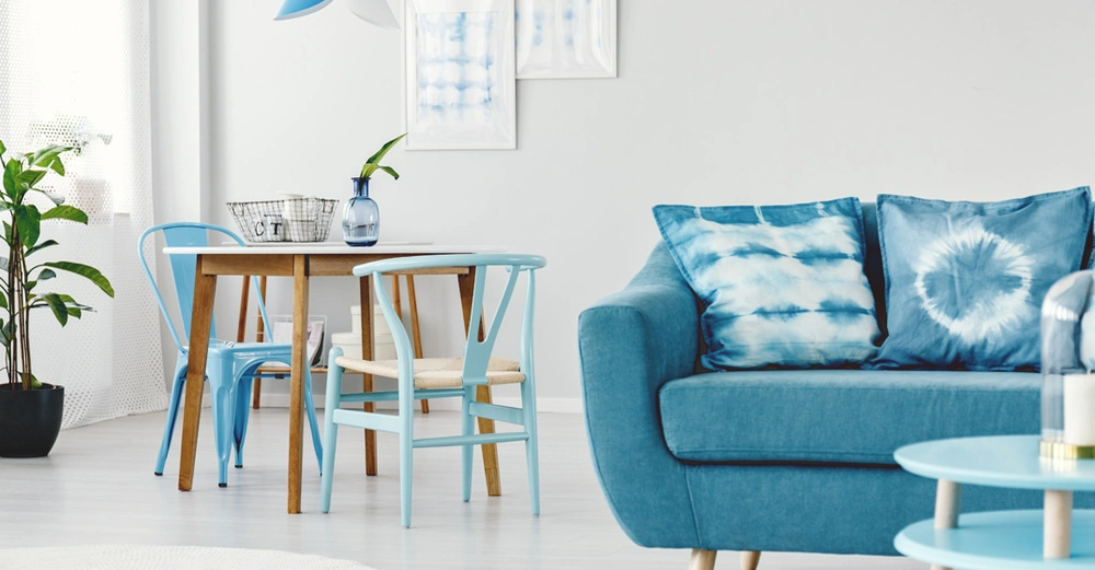 Blaues Sofa mit Kissen steht vor einem Holztisch mit hellblauen Stühlen Ein Metallkorb und eine Vase befinden sich auf dem Tisch Pflanzen und Wandbilder schmücken den hellen Raum