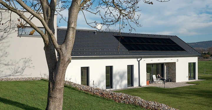Ein modernes weißes Haus hat schwarze Solarpaneele auf dem Dach zwei weiße Stühle stehen vor dem Haus auf einem Kiesweg umgeben von grüner Wiese und bedecktem Baum.