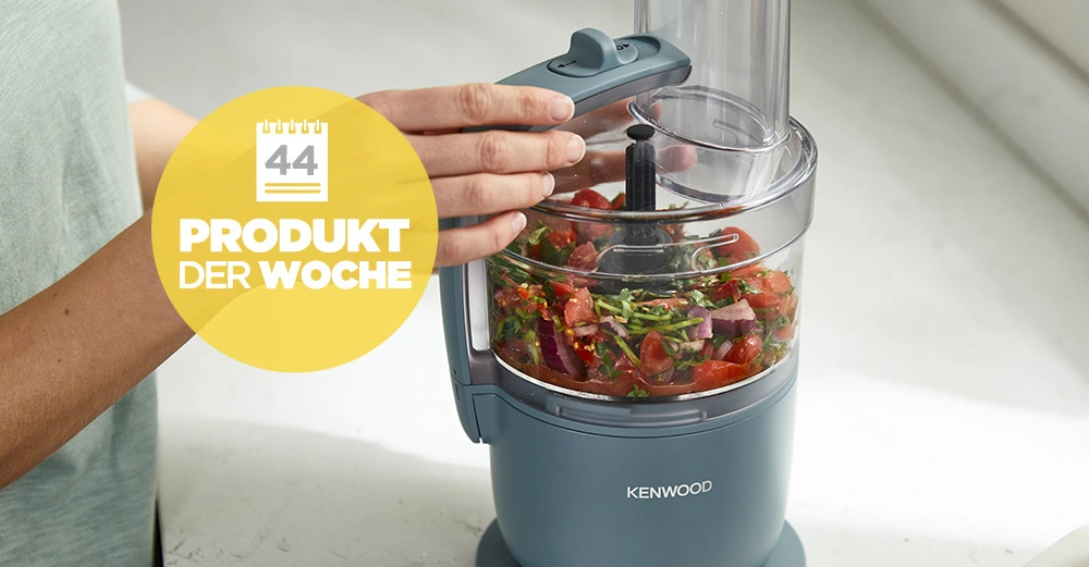 Eine Person bedient eine Kenwood-Küchenmaschine, die Gemüse zerkleinert. Die Küchenmaschine steht auf einer Küchenarbeitsplatte. Text: "44 PRODUKT DER WOCHE".