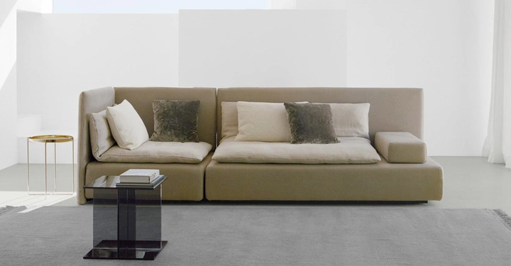 Beiges Sofa mit Kissen steht in schlichtem modernen Raum mit zwei Beistelltischen aus Glas und Metall vor weißen Wänden und einem grauen Teppich.