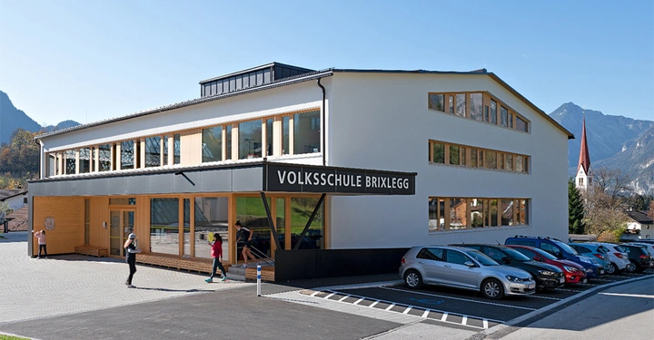 Galerie-Bild 2: Gebäude der Volksschule Brixlegg Menschen gehen hinein im Vordergrund parken Autos im Kontext von Alpengipfeln und einem Kirchturm im Hintergrund