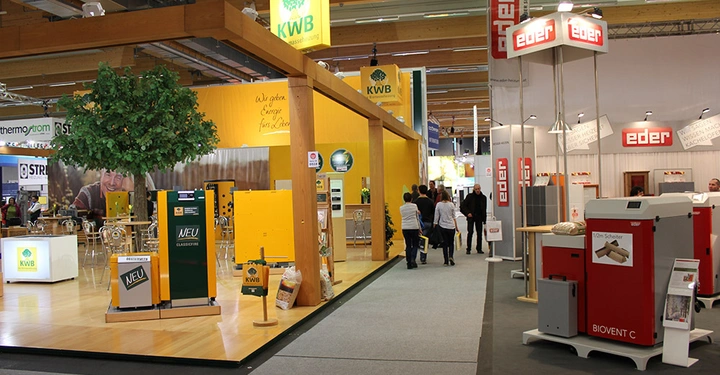 Galerie-Bild 5: Industrie-Ausstellungsbereich zeigt gelbe und rote Maschinen für Energie und Heizen unter Holzmasten. Besucher gehen herum. Text beinhaltet KWB Neu Wohnkomfort aus Biomasseheizung und Eder Biovent C und WIR SCHAFFEN WÄRME.