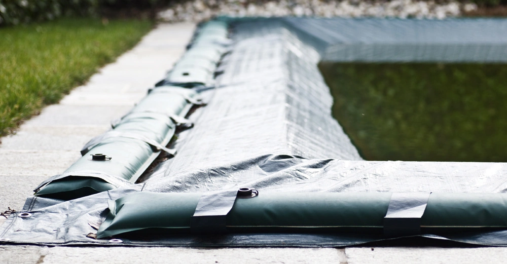 Abdeckung bedeckt ein rechteckiges Schwimmbecken auf einer Steinterrasse; grüne Sandsäcke halten die Abdeckung fest; umgeben von Rasenfläche und einer Hecke im Hintergrund rechts Kieselsteine.