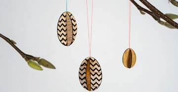 Eiförmige Holzornamente hängen an bunten Fäden von Zweigen. Die Ornamente haben Zickzack-Muster und die Zweige haben Knospen. Der Hintergrund ist einfarbig weiß.