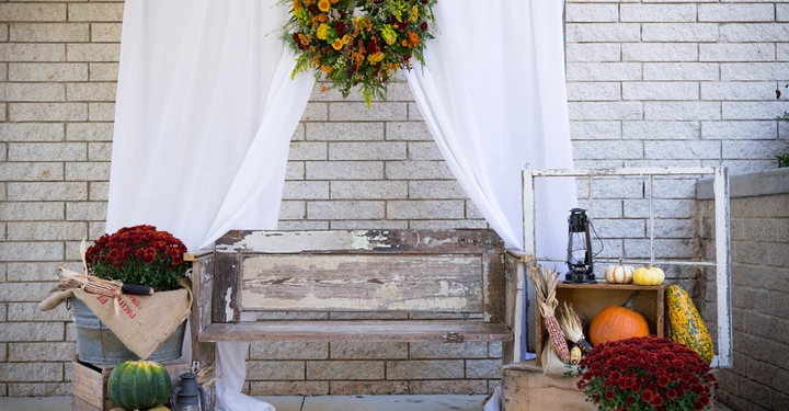 Galerie-Bild 10: Eine alte Holzbank steht vor einer Backsteinmauer mit einem weißen Vorhang im Hintergrund; dekoriert mit Kürbissen, herbstlichen Blumen und einem Kranz.