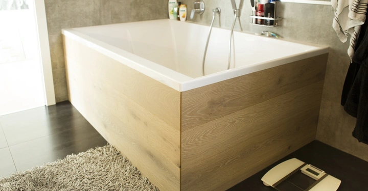 Galerie-Bild 3: Badewanne mit Holzverkleidung steht in modernem Badezimmer Teppich darunter Waage daneben Shampoos und Handtücher an der Wand befestigt großes Fenster im Hintergrund helle Fliesen und Wände